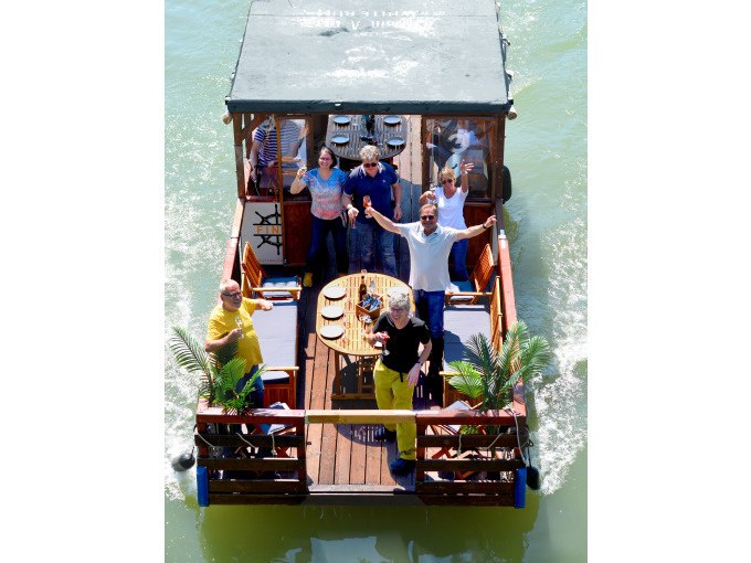 Eine Gruppe von Menschen steht auf einem Floßboot mit Tisch und Stühlen, während es auf einem Fluss fährt. Sie wirken fröhlich und entspannt., © Cool-Tours StattReisen Eine Gruppe von Menschen steht auf einem Floßboot mit Tisch und Stühlen, während es auf einem Fluss fährt. Sie wirken fröhlich und entspannt., © Cool-Tours StattReisen