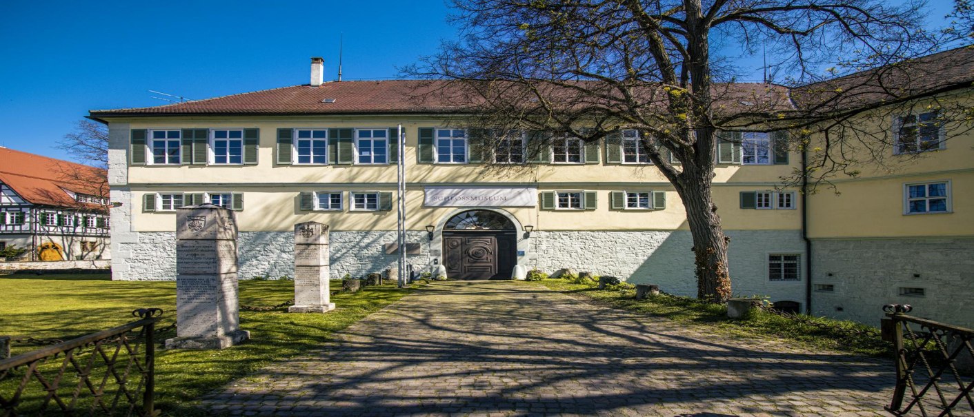 Schloss Kirchheim unter Teck: Historisches Gebäude mit gelber Fassade, Innenhof, Baum und Gedenksteinen. Blauer Himmel im Hintergrund., © SMG, Sarah Schmid