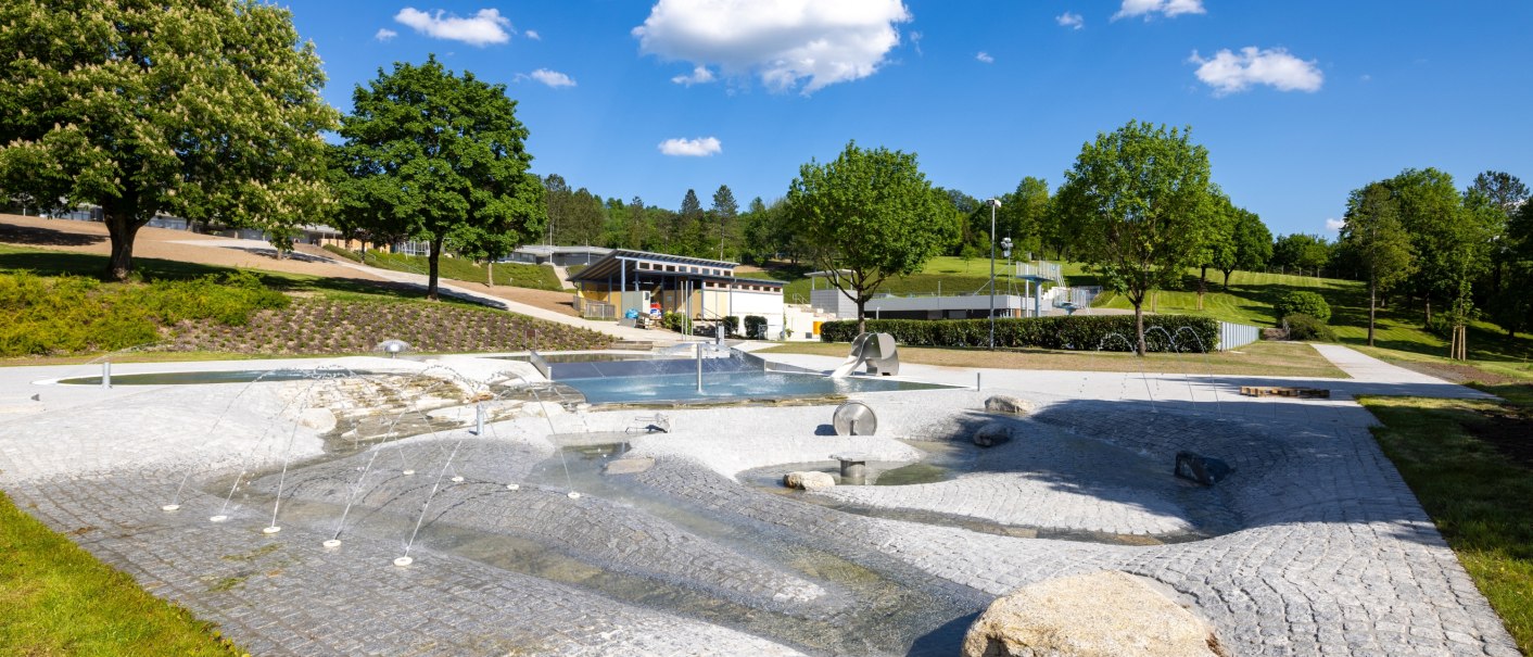 gepflasterter Wasserspielbereich mit Wasserfontänen sowie Wasserlauf mit Wasserrad, dahinter Plaschbecken mit kleiner Wasserrutsche in Elefantenform,  im Bildhintergrund Liegewiese und Servicegebäude, © Stadt Gaildorf