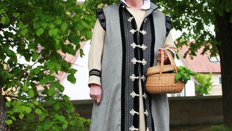 Eine Frau in historischer Kleidung h&auml;lt einen Korb und steht auf einem gepflasterten Weg unter einem Baum. Sie tr&auml;gt ein langes Gewand und eine wei&szlig;e Haube., &copy; Stadt Leonberg