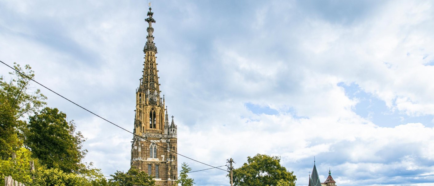 Gotischer Kirchturm in Esslingen ragt in den bewölkten Himmel. Umgeben von Bäumen und einem weiteren Turm im Hintergrund., © Stuttgart-Marketing GmbH, Sarah Schmid Gotischer Kirchturm in Esslingen ragt in den bewölkten Himmel. Umgeben von Bäumen und einem weiteren Turm im Hintergrund., © Stuttgart-Marketing GmbH, Sarah Schmid