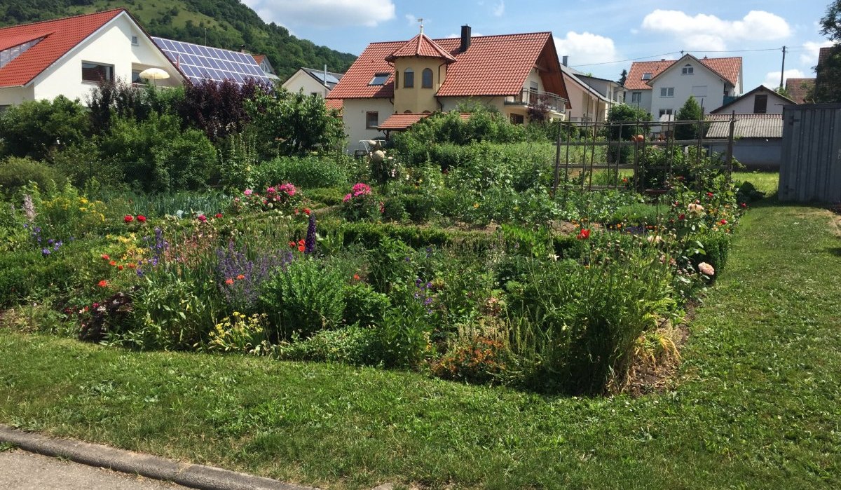 Ein blühender Vorgarten mit bunten Blumen und Sträuchern vor mehreren Häusern mit roten Dächern und Solarpanelen. Im Hintergrund sind grüne Hügel zu sehen., © www.pro-cycl.de Ein blühender Vorgarten mit bunten Blumen und Sträuchern vor mehreren Häusern mit roten Dächern und Solarpanelen. Im Hintergrund sind grüne Hügel zu sehen., © www.pro-cycl.de