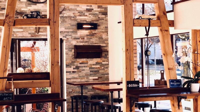 Gemütlicher Innenraum mit Holzbalken, hohen Tischen und Hockern. Dekorationen wie ein Modellfahrrad und eine große Uhr an der Steinwand., © Bady's Gemütlicher Innenraum mit Holzbalken, hohen Tischen und Hockern. Dekorationen wie ein Modellfahrrad und eine große Uhr an der Steinwand., © Bady's