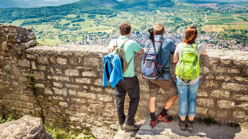 Drei Wanderer mit Rucks&auml;cken stehen auf einer Mauer und genie&szlig;en den Ausblick auf die gr&uuml;ne Landschaft von Hohenneuffen., &copy; hochgehberge