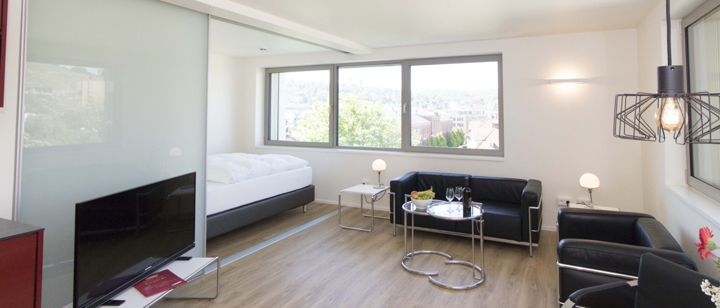 Modernes Apartment mit Holzboden, großem Fenster, schwarzem Sofa, Fernseher und Esstisch. Helle, minimalistische Einrichtung., © Büroma-Apart Apartmentvermietung GmbH Modernes Apartment mit Holzboden, großem Fenster, schwarzem Sofa, Fernseher und Esstisch. Helle, minimalistische Einrichtung., © Büroma-Apart Apartmentvermietung GmbH