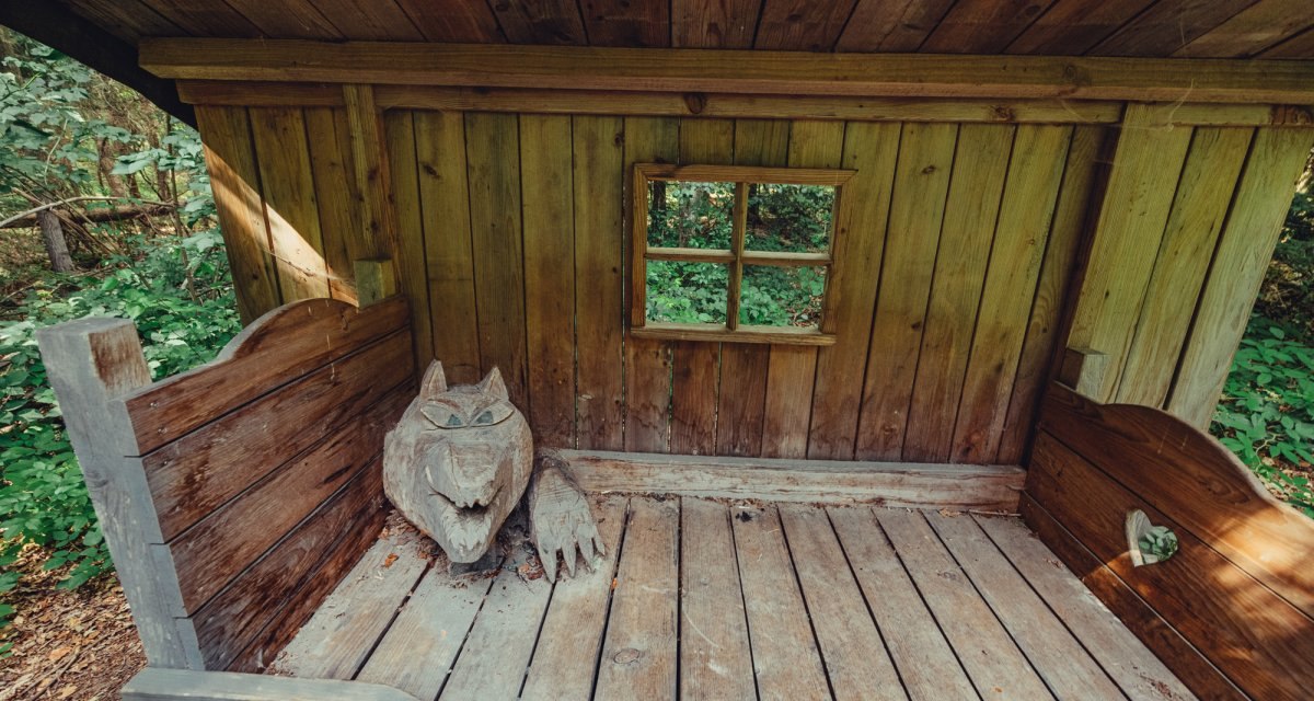 Eine Holzhütte im Märchenwald mit einer geschnitzten Wolfsskulptur auf einem Holzbett. Ein kleines Fenster zeigt den Wald im Hintergrund., © Tourismus Ostalb Eine Holzhütte im Märchenwald mit einer geschnitzten Wolfsskulptur auf einem Holzbett. Ein kleines Fenster zeigt den Wald im Hintergrund., © Tourismus Ostalb