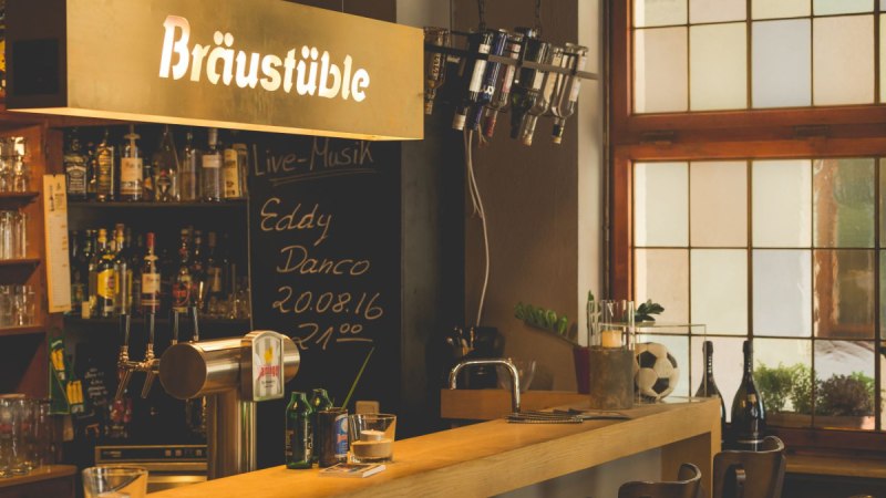 Gemütliche Bar mit Holztheke, Flaschenregal und Schild 'Bräustüble'. Schwarzes Brett kündigt Live-Musik an. Fenster im Hintergrund. Gemütliche Bar mit Holztheke, Flaschenregal und Schild 'Bräustüble'. Schwarzes Brett kündigt Live-Musik an. Fenster im Hintergrund.