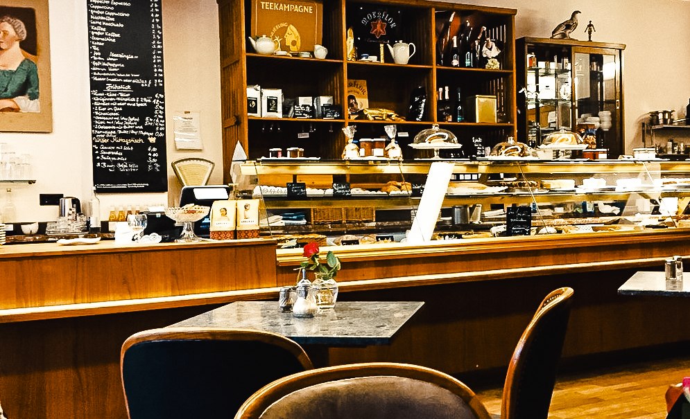 Ein gemütliches Café mit einer Theke voller Gebäck, Regalen mit Tee und Getränken. Tische mit Blumen und eine Getränkekarte an der Wand., © KoenigX