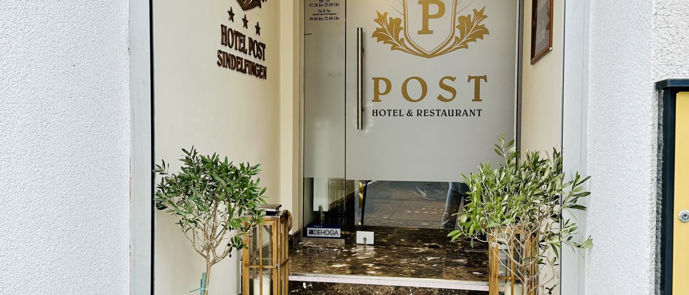Eingang zum POST Hotel & Restaurant mit Glastür, Marmorstufen und Pflanzen in Töpfen. An der Wand ist das Logo des Hotels zu sehen., © Hotel Post