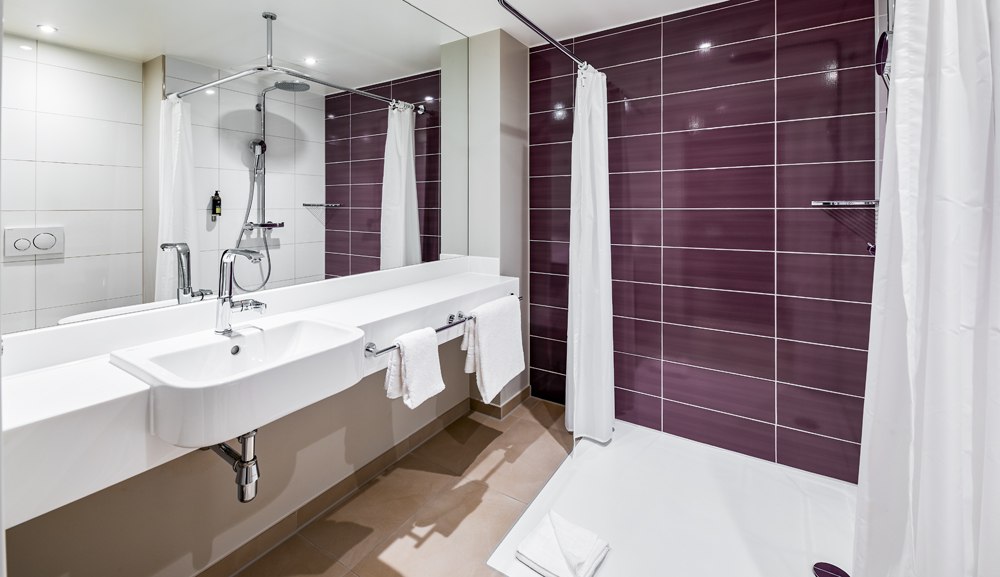 Modernes barrierefreies Badezimmer mit ebenerdiger Dusche, weißen und lila Fliesen, Waschbecken und Handtüchern., © Premier Inn
