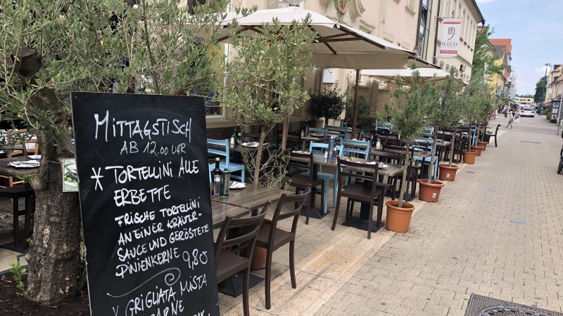 Ein Straßencafé mit Tischen und Stühlen unter Sonnenschirmen. Eine Menütafel bietet Tortellini und Fleischgerichte an. Olivenbäume in Töpfen säumen den Weg., © Antipasto Wein und mehr