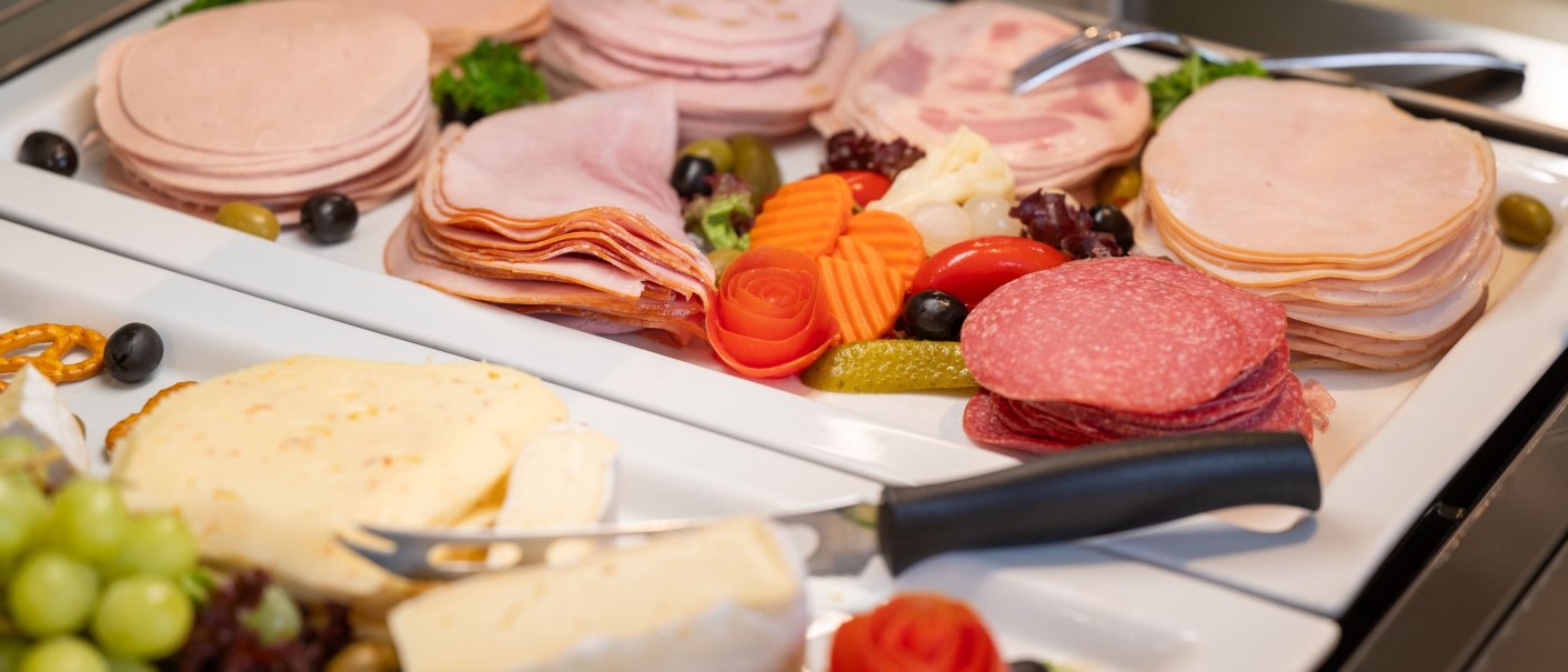 Ein Frühstücksbuffet mit verschiedenen Wurst- und Käsesorten, dekoriert mit Gemüse und Oliven auf weißen Platten., © Erikson Hotel Ein Frühstücksbuffet mit verschiedenen Wurst- und Käsesorten, dekoriert mit Gemüse und Oliven auf weißen Platten., © Erikson Hotel