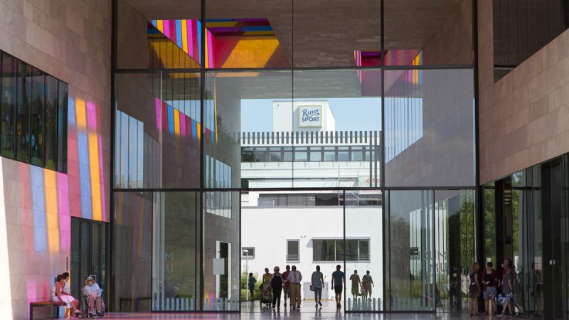 Glaspassage im Museum Ritter mit bunten Lichtreflexionen. Menschen gehen durch die Passage, im Hintergrund ist das Ritter Sport Logo sichtbar., &copy; Region Stuttgart