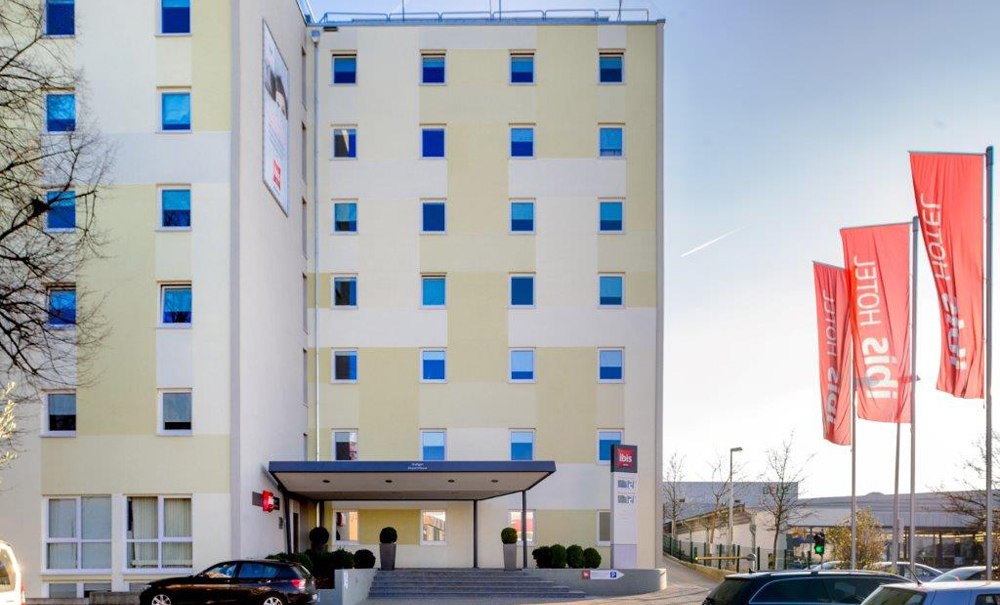 Das Ibis Hotel Stuttgart Airport Messe mit roten Fahnen und geparkten Autos davor. Das Gebäude hat mehrere Stockwerke und eine helle Fassade., © Ibis Stuttgart Airport Messe Das Ibis Hotel Stuttgart Airport Messe mit roten Fahnen und geparkten Autos davor. Das Gebäude hat mehrere Stockwerke und eine helle Fassade., © Ibis Stuttgart Airport Messe