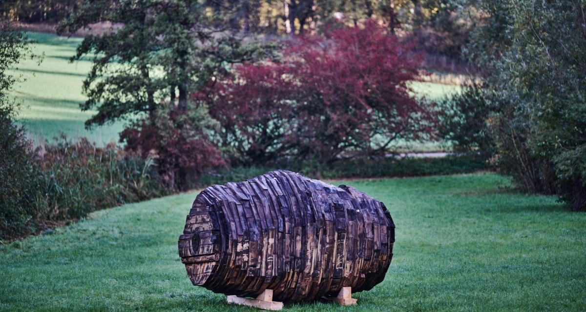 Holzskulptur in Fassform auf grüner Wiese, umgeben von Bäumen und Sträuchern. Die Skulptur besteht aus dunklen, geschichteten Holzstücken., © Natur.Nah. Schönbuch & Heckengäu Holzskulptur in Fassform auf grüner Wiese, umgeben von Bäumen und Sträuchern. Die Skulptur besteht aus dunklen, geschichteten Holzstücken., © Natur.Nah. Schönbuch & Heckengäu