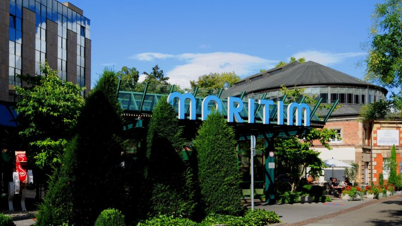 Au&szlig;enansicht des Maritim Hotels mit markantem Glasdach und umgeben von B&auml;umen und Pflanzen. Ein Weg f&uuml;hrt am Geb&auml;ude entlang., &copy; Maritim Hotelgesellschaft mbH