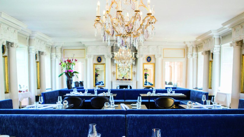 Elegantes Restaurant mit Kronleuchter, blauen Sitzbänken und weißen Säulen. Blumenarrangements und gedeckte Tische schaffen eine luxuriöse Atmosphäre., © Speisemeisterei