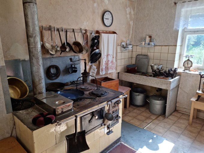 Eine nostalgische Küche mit Holzofen, Pfannen und Töpfen an der Wand, einer Spüle und einer Waage auf der Fensterbank. Sonnenlicht fällt durch das Fenster., © SwabianTravel Eine nostalgische Küche mit Holzofen, Pfannen und Töpfen an der Wand, einer Spüle und einer Waage auf der Fensterbank. Sonnenlicht fällt durch das Fenster., © SwabianTravel