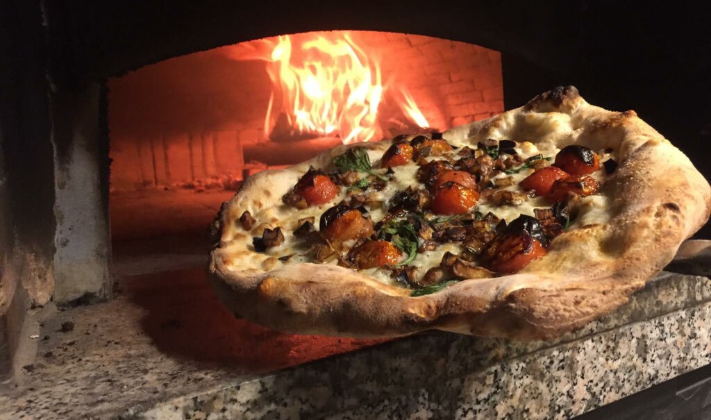 Eine frisch gebackene Pizza mit Tomaten und Kr&auml;utern wird aus einem hei&szlig;en Holzofen geholt. Der Ofen ist aus Stein und das Feuer lodert., &copy; DaCarmine