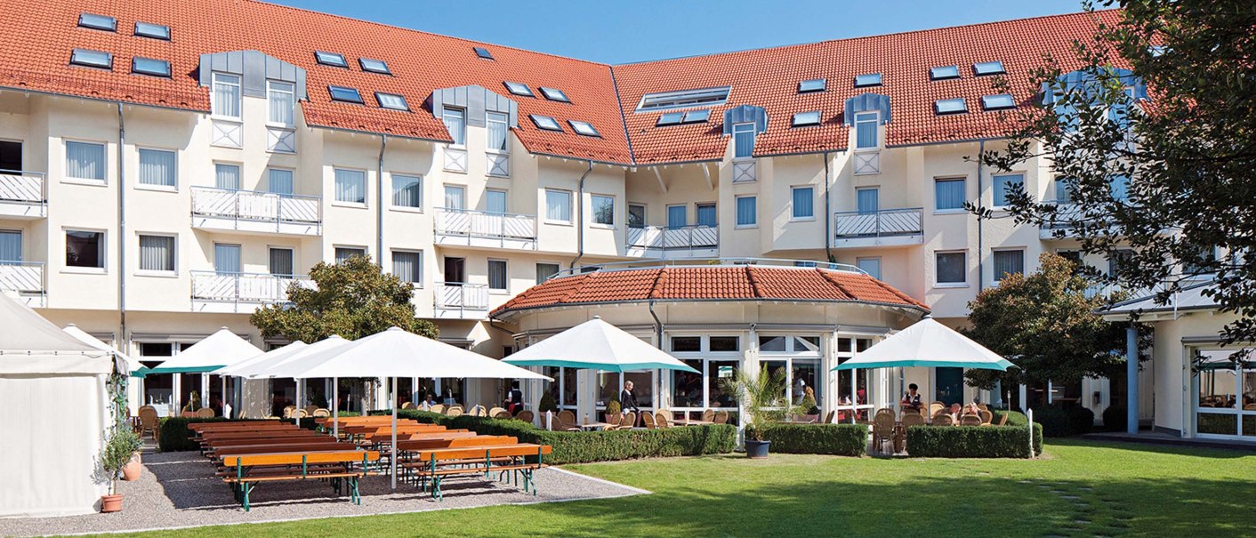 Biergarten mit Holzbänken und weißen Sonnenschirmen vor einem großen Gebäude mit roten Dächern und vielen Fenstern., © Seminaris Hotel Bad Boll Biergarten mit Holzbänken und weißen Sonnenschirmen vor einem großen Gebäude mit roten Dächern und vielen Fenstern., © Seminaris Hotel Bad Boll