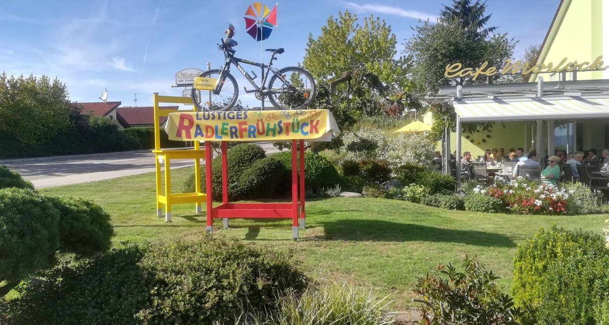 Café Morlock mit Terrasse, Menschen sitzen draußen. Im Vordergrund ein buntes Schild mit Fahrrädern und 'Lustiges Radlerfrühstück'., © Kulturamt Plochingen Café Morlock mit Terrasse, Menschen sitzen draußen. Im Vordergrund ein buntes Schild mit Fahrrädern und 'Lustiges Radlerfrühstück'., © Kulturamt Plochingen