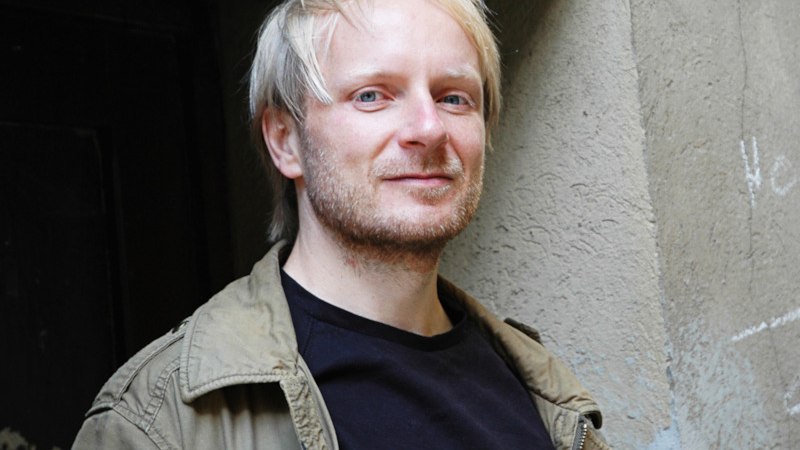 Ein Mann mit blonden Haaren und Bart steht lächelnd vor einer grauen Wand. Er trägt eine olivgrüne Jacke und ein schwarzes T-Shirt., © Kulturamt der Stadt Esslingen am Neckar
