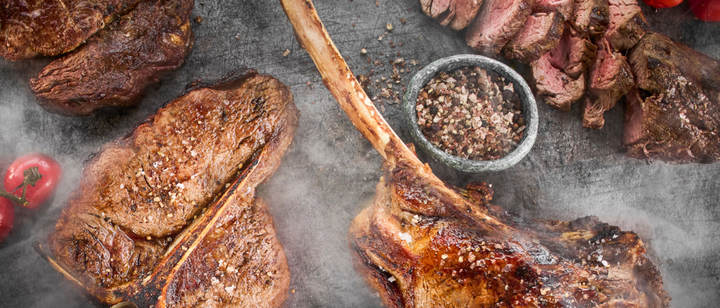 Gegrillte Steaks auf einem Tisch, umgeben von Gewürzen und Tomaten. Die Steaks sind saftig und mit Salz bestreut, daneben eine Schale mit Pfefferkörnern., © MAREDO