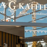 Eingang des Glora Kaffeehauses mit dekorativem Schriftzug auf der Glasfront. Im Hintergrund sind Baukräne zu sehen., © SMG, Sarah Schmid Eingang des Glora Kaffeehauses mit dekorativem Schriftzug auf der Glasfront. Im Hintergrund sind Baukräne zu sehen., © SMG, Sarah Schmid