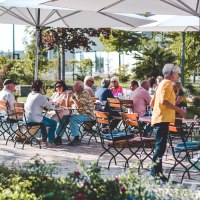 Menschen sitzen unter großen Sonnenschirmen an Holztischen im Freien, umgeben von grüner Vegetation und Blumen., © Wilhelm Kern GmbH Menschen sitzen unter großen Sonnenschirmen an Holztischen im Freien, umgeben von grüner Vegetation und Blumen., © Wilhelm Kern GmbH