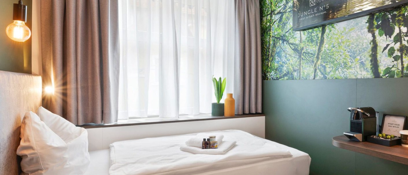 Modernes Einzelzimmer mit Bett, großem Fenster, Dschungel-Wandbild und Kaffeemaschine. Dezente Beleuchtung und stilvolle Dekoration., © JUNGLE N°5 GmbH