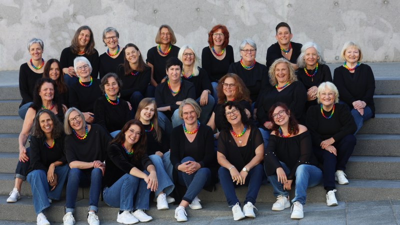 Eine fr&ouml;hliche Frauengruppe sitzt auf Treppenstufen, tr&auml;gt schwarze Kleidung und bunte Halsketten. Sie l&auml;cheln in die Kamera., &copy; metSingers