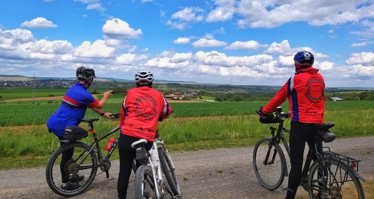 Drei Radfahrer in bunter Sportkleidung stehen auf einem Feldweg und schauen in die weite, grüne Landschaft unter einem blauen Himmel mit Wolken., © Land der 1000 Hügel - Kraichgau-Stromberg Drei Radfahrer in bunter Sportkleidung stehen auf einem Feldweg und schauen in die weite, grüne Landschaft unter einem blauen Himmel mit Wolken., © Land der 1000 Hügel - Kraichgau-Stromberg