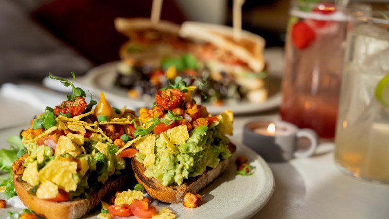 Avocado-Toast mit Tomaten und Kräutern, im Hintergrund ein Sandwich und Getränke. Ein Teelicht sorgt für gemütliche Atmosphäre., © Veganes Gold®