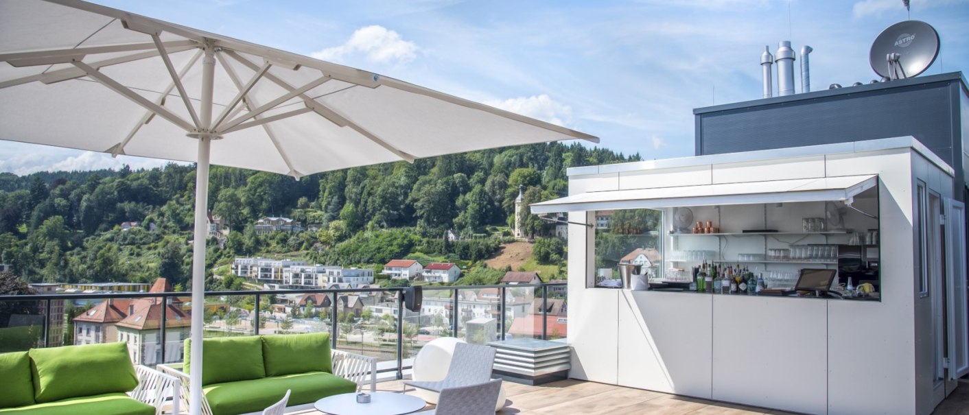 Moderne Dachterrassenbar mit Sonnenschirm, grünen Sofas und Blick auf eine grüne Hügellandschaft., © Hotel am Remspark Moderne Dachterrassenbar mit Sonnenschirm, grünen Sofas und Blick auf eine grüne Hügellandschaft., © Hotel am Remspark