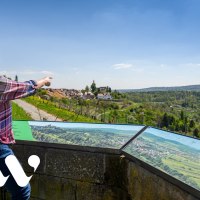 Eine Person zeigt auf eine malerische Landschaft mit Hügeln und einem Dorf. Im Vordergrund sind Informationstafeln zu sehen.