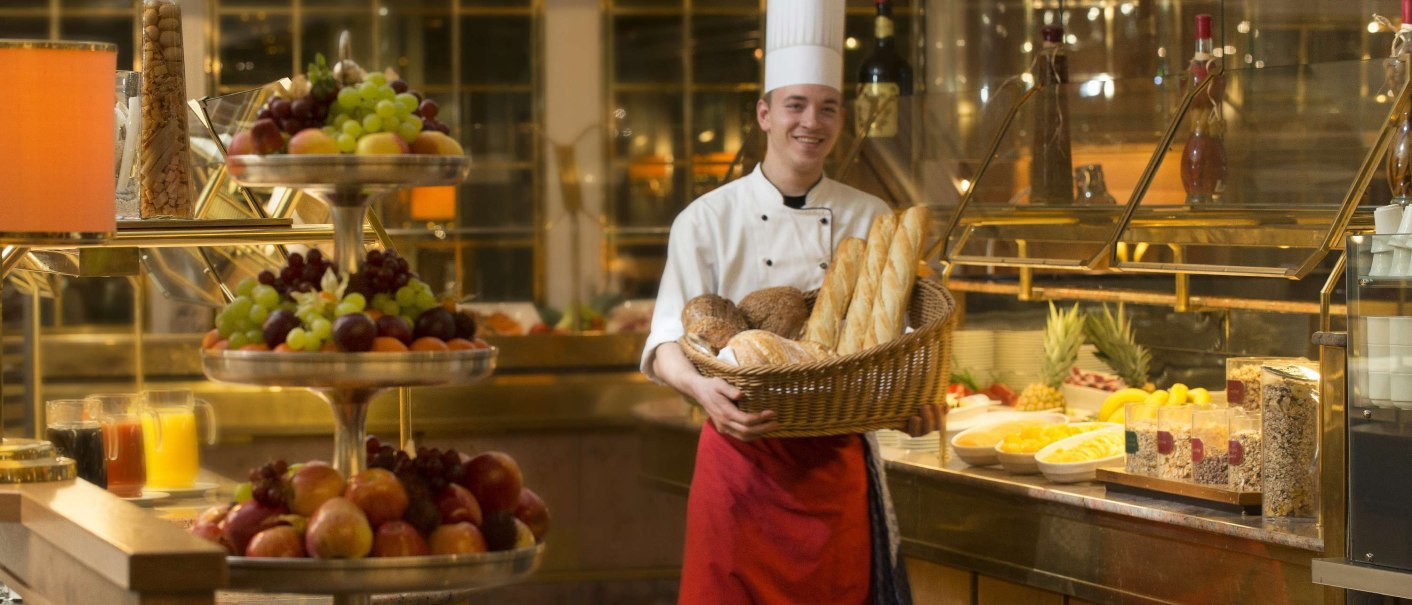 Ein Koch in Uniform hält einen Korb mit Brot vor einem reichhaltigen Frühstücksbuffet mit Obst, Saft und Müsli., © Maritim Hotelgesellschaft mbH