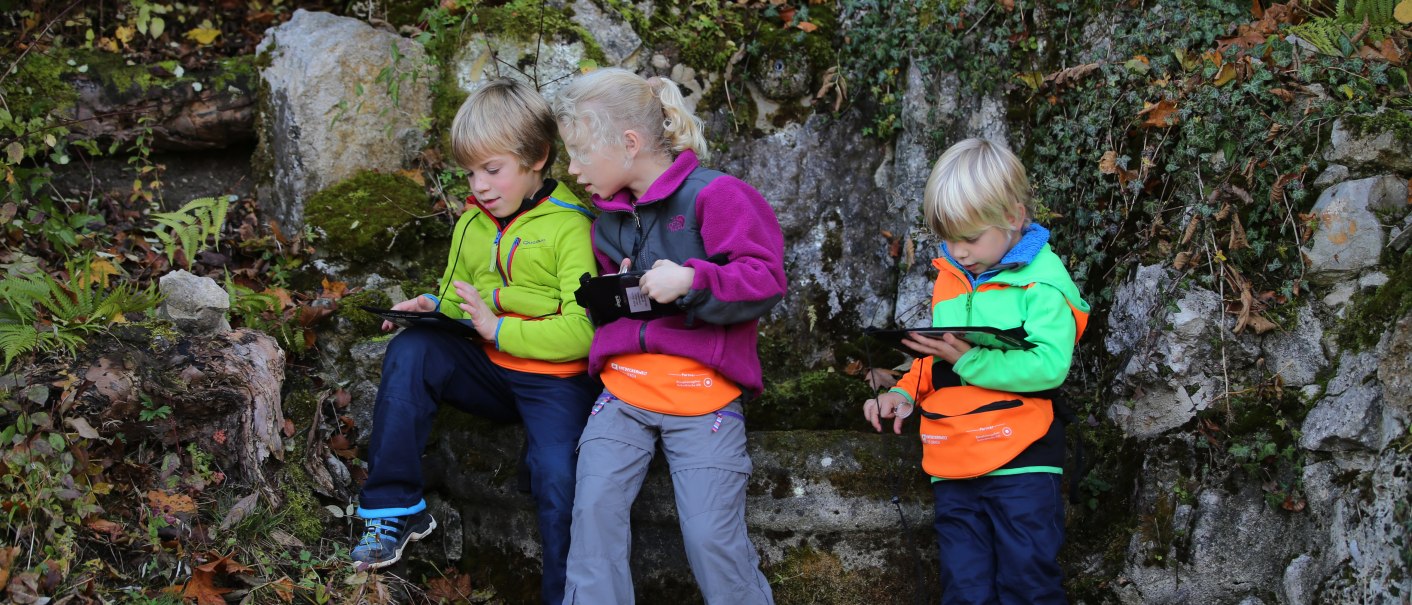 Drei Kinder in bunter Outdoor-Kleidung sitzen im Wald und schauen auf Tablets. Sie sind von Pflanzen und Felsen umgeben., © Bad Urach Tourismus