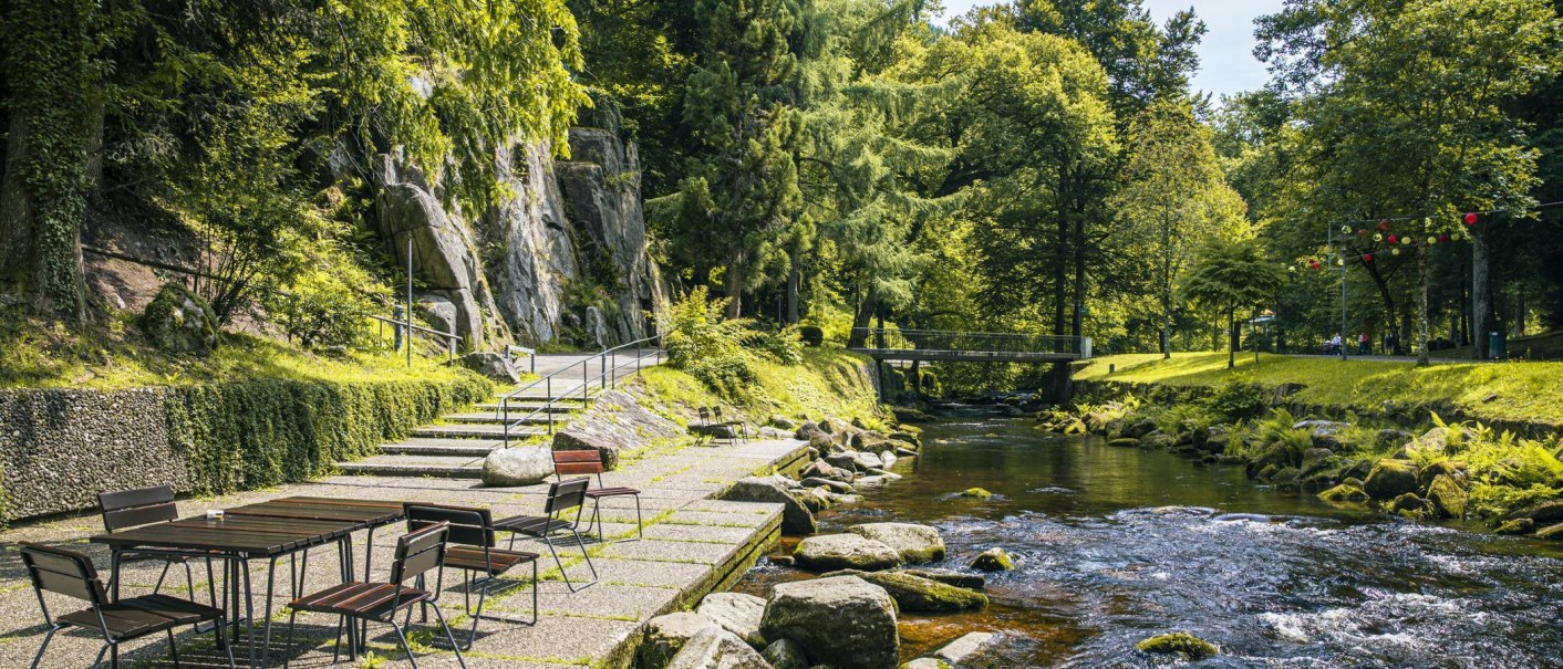 Ein ruhiger Fluss flie&szlig;t durch einen gr&uuml;nen Park mit B&auml;umen und einem Steg. Links stehen Tische und St&uuml;hle auf einer Terrasse., &copy; Stuttgart-Marketing GmbH, Sarah Schmid