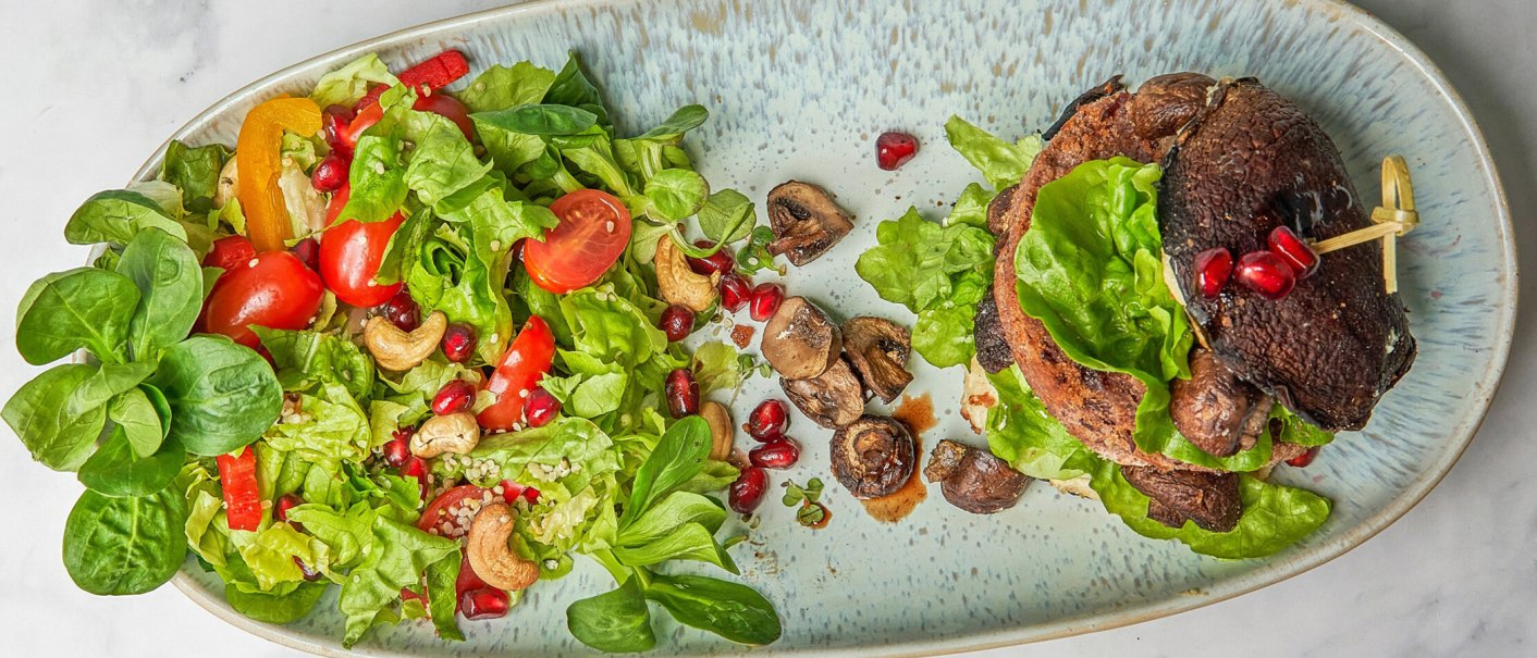 Ein Teller mit Salat aus grünem Blattgemüse, Tomaten, Paprika, Cashewnüssen und Granatapfelkernen. Daneben ein Burger mit Pilzen., © SMG Ein Teller mit Salat aus grünem Blattgemüse, Tomaten, Paprika, Cashewnüssen und Granatapfelkernen. Daneben ein Burger mit Pilzen., © SMG