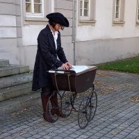 Eine Person in historischer Kleidung steht neben einem antiken Kinderwagen auf einem gepflasterten Weg vor einem Geb&auml;ude mit wei&szlig;en Fenstern., &copy; Margit Sch&auml;fer