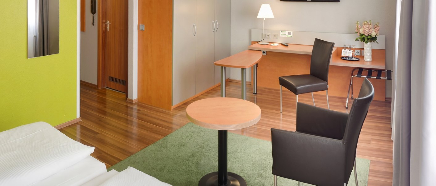 Modernes Hotelzimmer mit Doppelbett, Schreibtisch, Stühlen, Flachbildfernseher und grünem Teppich auf Holzboden., © attimo Hotel Stuttgart
