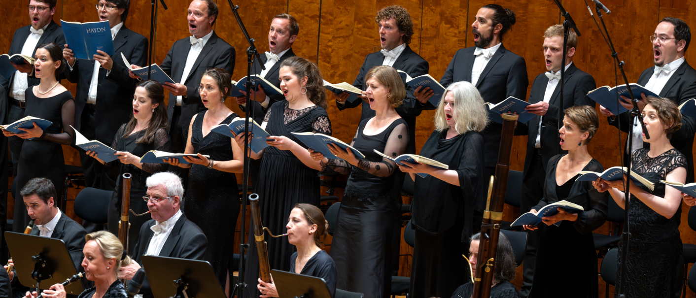 Ein Chor und Orchester in eleganter Kleidung f&uuml;hren ein Werk auf. Die S&auml;nger halten Notenbl&auml;tter und singen mit Ausdruck., &copy; Holger Schneider