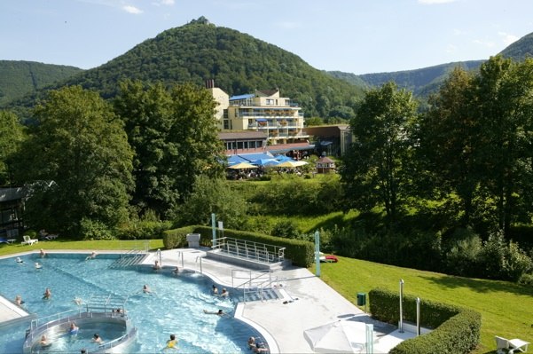 Hotel mit Pool vor bewaldeten Hügeln, umgeben von grüner Landschaft und Bäumen, unter blauem Himmel., © ©hotel-graf-eberhard.de