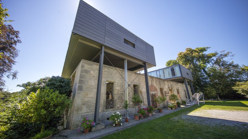 Modernes Gebäude mit alten Steinmauern und Glasstruktur, umgeben von Pflanzen und Bäumen unter blauem Himmel., © Region Stuttgart Modernes Gebäude mit alten Steinmauern und Glasstruktur, umgeben von Pflanzen und Bäumen unter blauem Himmel., © Region Stuttgart