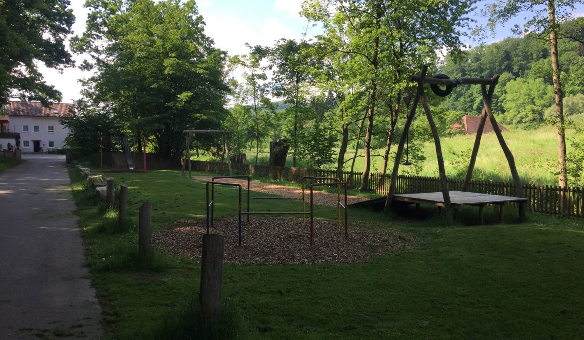 Ein ruhiger Spielplatz mit Schaukeln, Rutsche und Klettergerüst, umgeben von Bäumen und Wiese. Ein Weg führt an einem Gebäude vorbei., © www.pro-cycl.de Ein ruhiger Spielplatz mit Schaukeln, Rutsche und Klettergerüst, umgeben von Bäumen und Wiese. Ein Weg führt an einem Gebäude vorbei., © www.pro-cycl.de