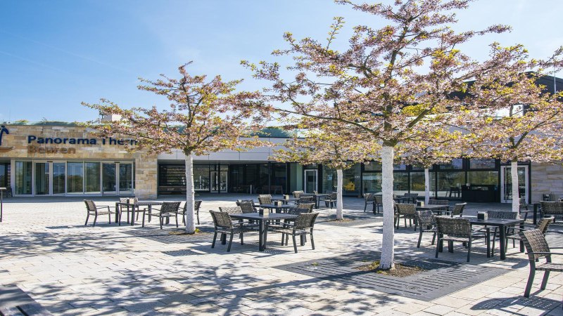 Haupteingang der Panorama Therme Beuren mit blühenden Bäumen und Bistro-Tischen im Freien., © SMG, Sarah Schmid Haupteingang der Panorama Therme Beuren mit blühenden Bäumen und Bistro-Tischen im Freien., © SMG, Sarah Schmid