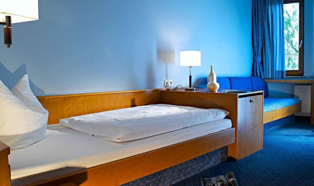 Ein gemütliches Einzelzimmer mit blauem Teppich, Einzelbett, Sofa und Fenster mit blauen Vorhängen. Eine Lampe und Zeitschrift sind sichtbar., © Nödinger Gastro GmbH