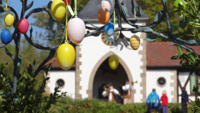Bunte Ostereier h&auml;ngen an einem Baum, im Hintergrund ein Geb&auml;ude mit Menschen. Fr&uuml;hlingshafte Szene mit festlicher Dekoration., &copy; Erlebnispark Tripsdrill