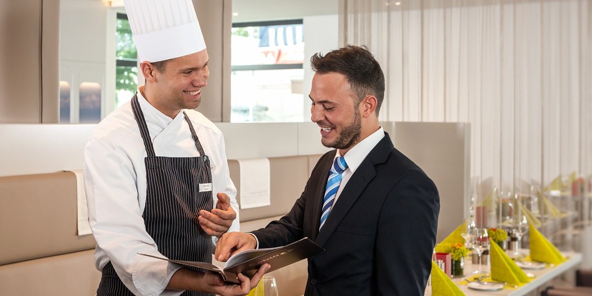 Ein Koch in Uniform und ein Mann im Anzug lächeln und betrachten ein Buch in einem eleganten Restaurant., © Essential by Dorint