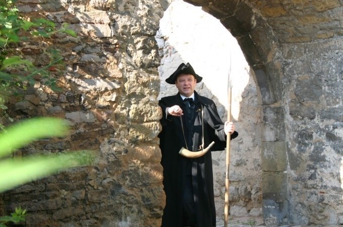Ein Mann in historischer Kleidung mit Horn und Stab steht unter einem steinernen Torbogen und zeigt auf etwas., © WTM GmbH Waiblingen